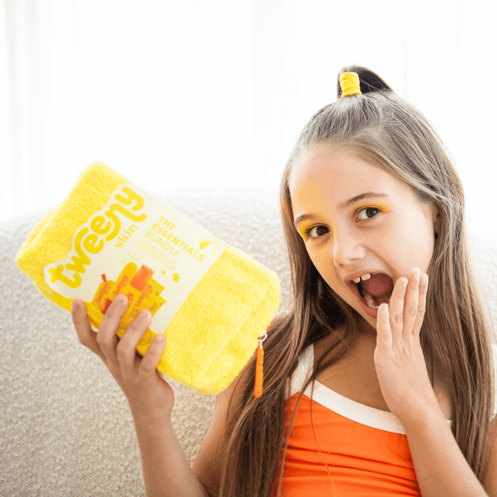 Excited tween girl holding the Tweeny Skin Essentials Bundle — a vibrant yellow skincare kit designed for tweens and teens. She’s wearing an orange outfit and matching yellow eye makeup, highlighting the fun, age-appropriate vibe of the brand. The Essentials Bundle includes gentle, fragrance-free products perfect for sensitive young skin, making it the ultimate starter set for building confident skincare habits.