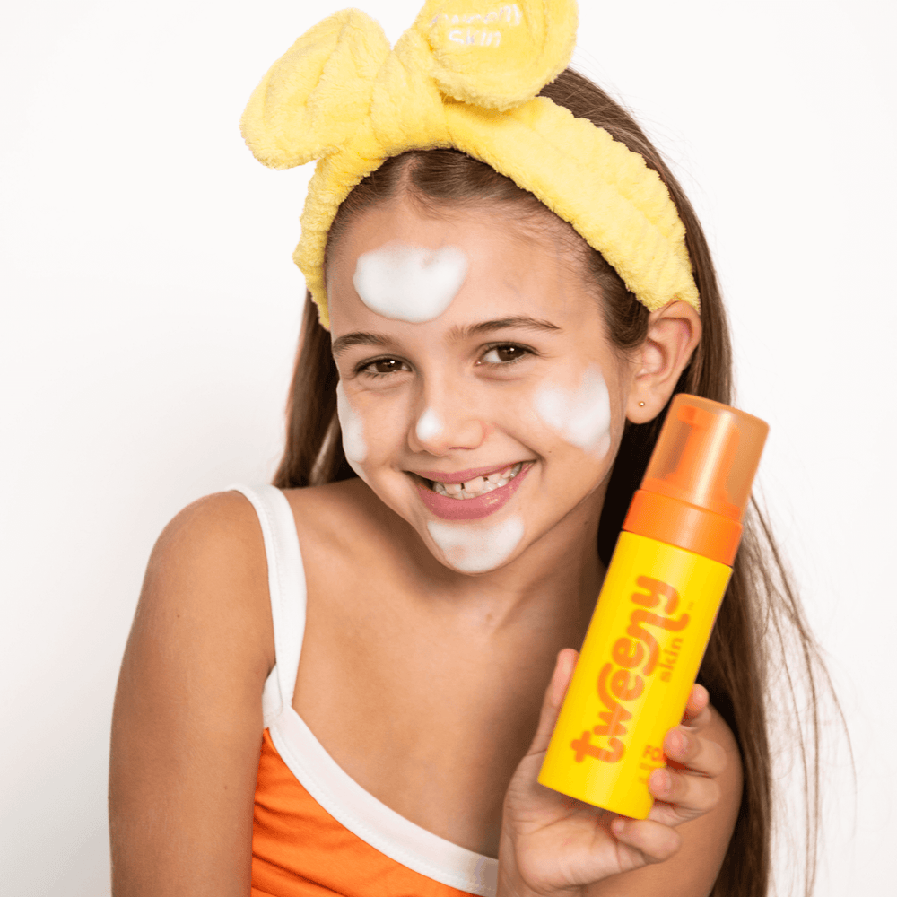 Smiling tween girl wearing a yellow Tweeny Skin Fluffy headband and orange top, holding Tweeny Skin FOAM-O Gentle Cleanser with foam on her face.