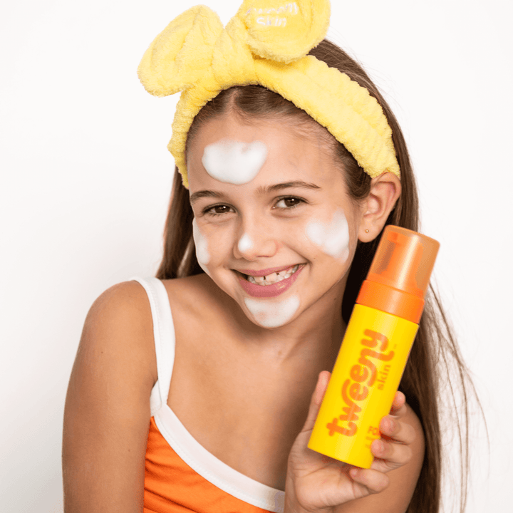 Smiling tween girl wearing a yellow Tweeny Skin Fluffy headband and orange top, holding Tweeny Skin FOAM-O Gentle Cleanser with foam on her face.