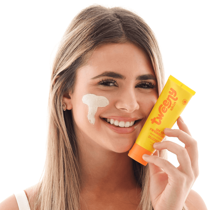 Smiling teen girl holding a tube of Tweeny Skin SPF 50 TweenScreen sunscreen, with a visible swipe of the product applied to her cheek.