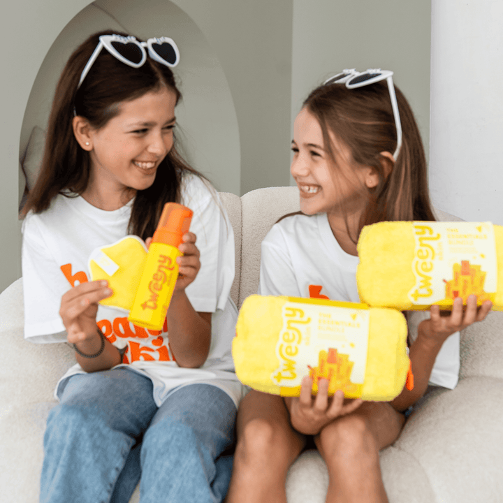 Two smiling tween girls sitting on a couch, laughing and holding the Tweeny Skin Essentials Bundle. One girl holds the yellow towel bag and FOAM-O Gentle Cleanser, while the other holds the full bundle set. Both wear matching white Tweeny Skin tees and heart-shaped sunglasses, capturing the fun and confidence the skincare range brings to tweens. Perfect visual for showcasing skincare made just for young, sensitive skin.